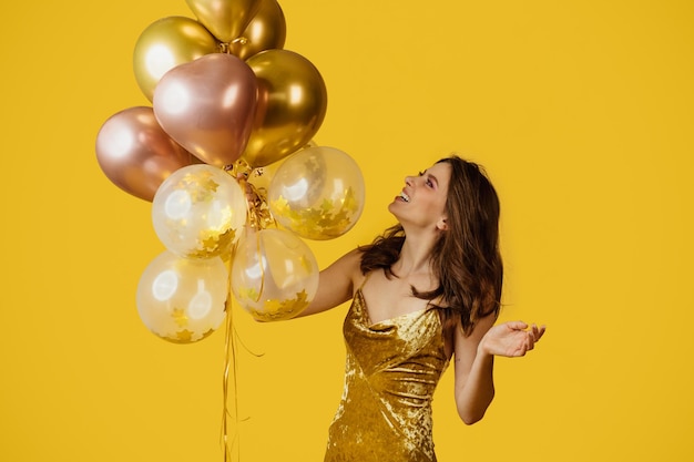 Holiday celebration concept Excited woman in festive dress holding bunch of balloons on yellow studio background