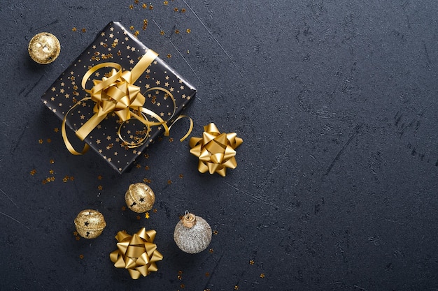 Holiday gift box or present with ribbon, golden confetti and gold baubles on black background. Magic christmas greeting card. Christmas Decoration. Border design. Mock up. Top view.