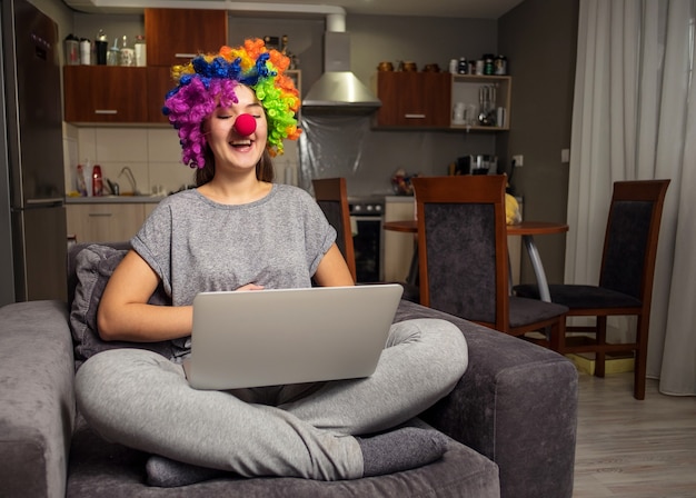 Holiday greetings online, young woman communicates in social networks with laptop, girl sitting at home in chair with laptop, clown with laptop at home, laugh