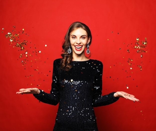 Holiday and party concept beautiful young woman wearing evening dress standing under confetti rain over red background