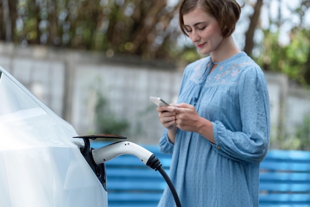 Holiday road trip vacation by the green countryside nature with beautiful young woman checking battery status from smartphone while recharging electric vehicleEcofriendly travel wit EV carPerpetual