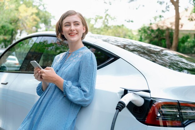 Holiday road trip vacation by the green countryside nature with beautiful young woman checking battery status from smartphone while recharging electric vehicleEcofriendly travel wit EV carPerpetual