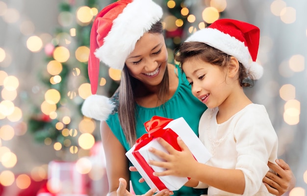 holidays, celebration, family and people concept - happy mother and little girl in santa helper hats with gift box over living room and christmas tree background