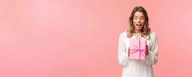 Holidays celebration and women concept Portrait of surprised charming young blond girl receive surprise gift holding present in pink box look at it amused curious whats inside studio background