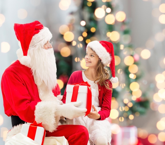 holidays, childhood and people concept - smiling little girl with santa claus and gifts over christmas tree lights lights background