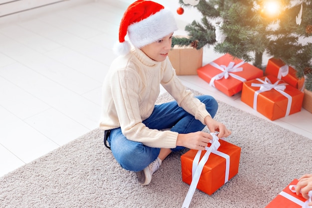 Holidays, christmas, childhood and people concept - smiling happy teen boy in santa hat opens gift box over christmas tree background.