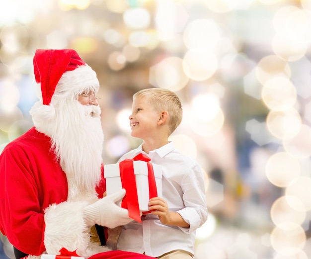 holidays, christmas, childhood and people concept - smiling little boy with santa claus and gifts over lights background