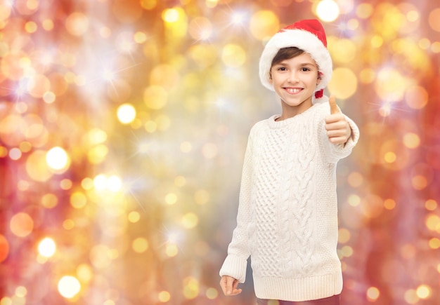 holidays, gesture, christmas, childhood and people concept - smiling happy boy in santa hat showing thumbs up over lights background