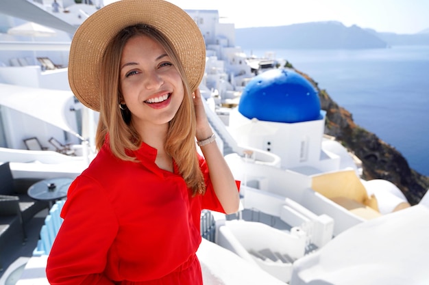 Holidays in Greece Closeup of smiling girl in Oia Santorini Relaxed woman with red dress and hat looking at camera and posing in Santorini with blue dome church in the background