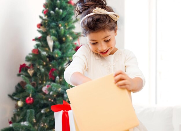 holidays, presents, childhood and people concept - smiling little girl with gift box over living room and christmas thee background