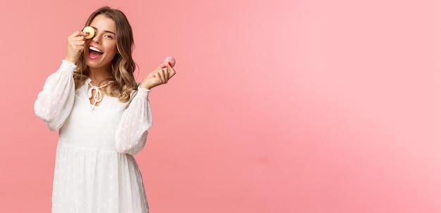 Holidays spring and party concept Portrait of carefree tender and feminine girl in white dress holding macarons dessert over eye feeling cheerful eating sweets tasty desserts pink background