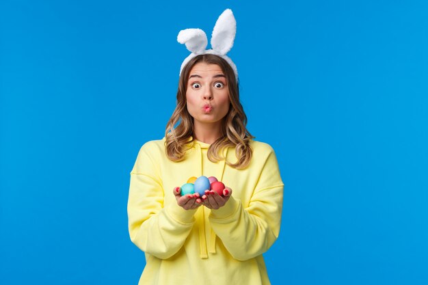 Holidays, traditions and celebration concept. Silly cute caucasian blond girl present you Easter eggs, painted it for holiday, show mwah kiss expression and wear rabbit ears, on a blue wall