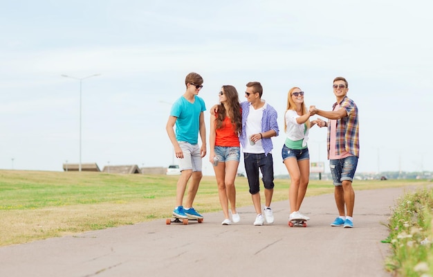 holidays, vacation, love and friendship concept - group of smiling teenagers walking and riding on skateboards outdoors