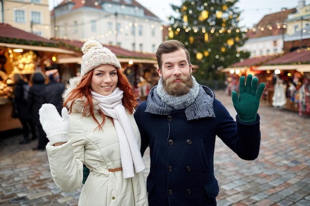 holidays, winter, christmas, gesture and people concept - happy couple of tourists in warm clothes waving hands in old town