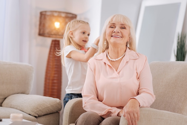 Home beauty salon. Cheerful positive aged woman sitting on the sofa and smiling while having her hairstyle made