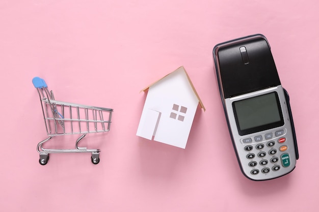 Home buying concept Mini house payment terminal and shopping trolley on pink blue background Top view