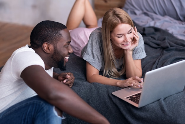 Home coziness. Young international delighted couple using laptop while lying in bedroom and smiling.