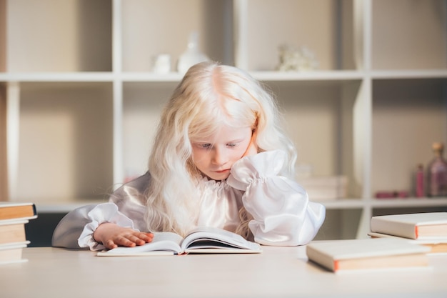 Home education. Child development. Pandemic self-isolation. Tired smart blonde albino little girl in white blouse reading book sitting at desk.