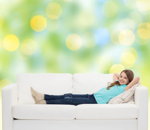 home, leisure, people and happiness concept - smiling little girl lying on sofa over green lights background