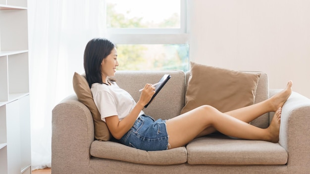 Home lifestyle concept Young woman lying on couch and using tablet to surf social media at home