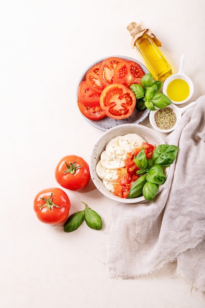 Home made healthy meal concept: cherry tomatoes, mozzarella balls, spices, olive oil and fresh basil. Mozzarella salad over white texture surface. Top view, flat lay