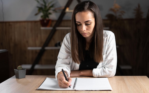 Home office brunette girl writes something with a pen in a notebook lying on a light table Adult female student studying at home remotely doing homework Online education concept