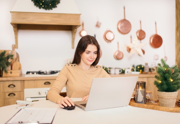 Home office concept. A young freelance woman working on a laptop at home in a bright kitchen in winter.