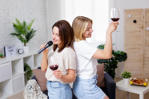 Home party - portrait of two beautiful girls having fun together, drinking wine, dancing and singing karaoke in cozy living room