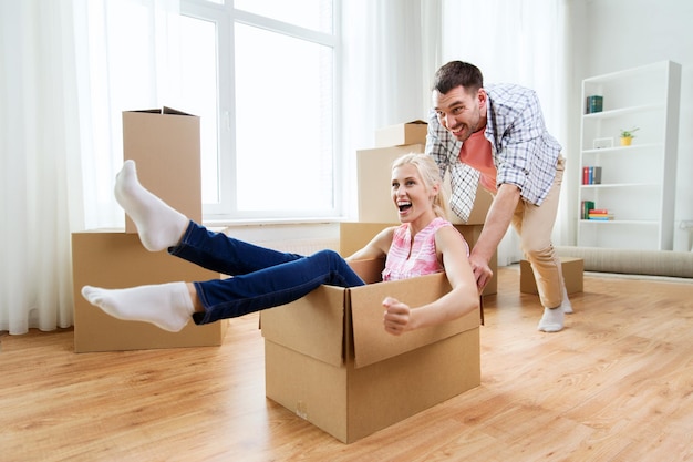 home, people, moving and real estate concept - happy couple having fun and riding in cardboard boxes at new home