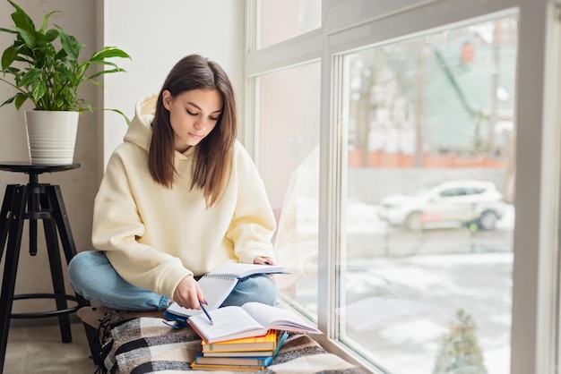 Home schooling A teenager girl a student sits on a windowsill with a bunch of books and writes homework Distance learning preparation for exams Soft selective focus