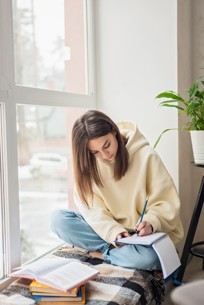 Home schooling A teenager girl a student sits on a windowsill with a bunch of books and writes homework Distance learning preparation for exams Soft selective focus