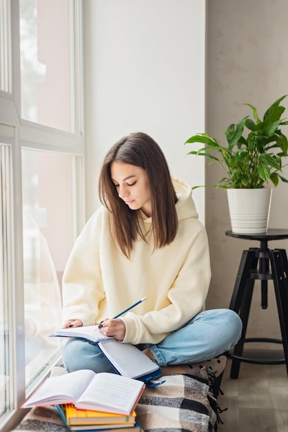 Home schooling A teenager girl a student sits on a windowsill with a bunch of books and writes homework Distance learning preparation for exams Soft selective focus