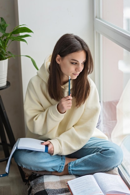 Home schooling A teenager girl a student sits on a windowsill with a bunch of books and writes homework Distance learning preparation for exams Soft selective focus