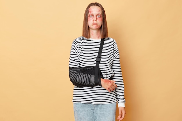 Home violence Trauma after car accident Unhappy disabled young girl with scratches and bruises on her face wearing striped shirt standing isolated over beige background