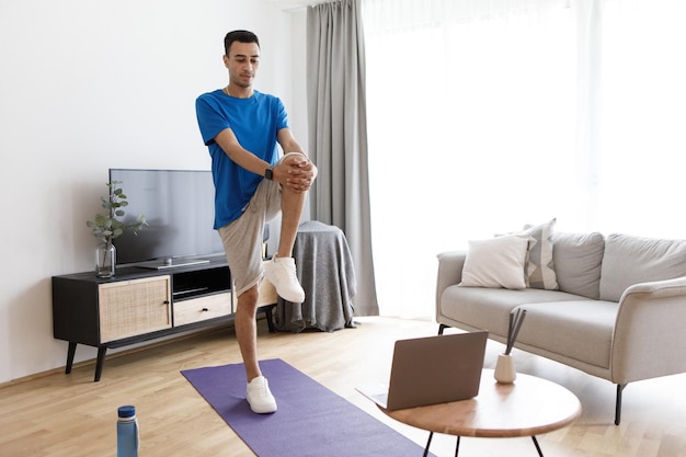 Home workout routine Young arab man stretching and doing standing kneetochest exercise using laptop computer