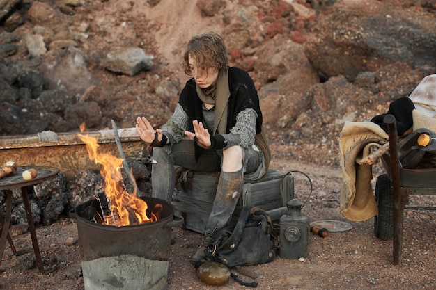 Homeless woman in torn clothes warming up near the fire outdoors among the rocks