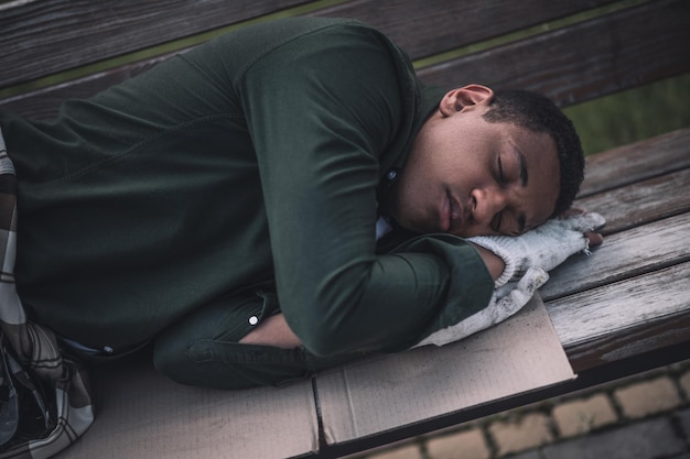 Homelessness. Dark-skinned young man holding folded arms under head sleeping on cardboard on bench outdoors during day