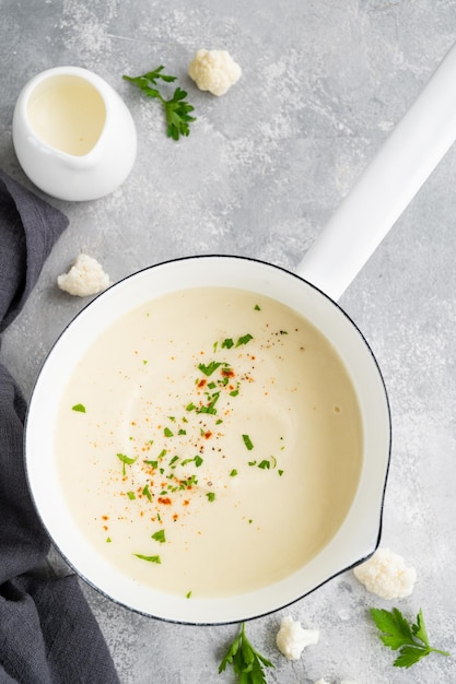 Homemade cauliflower soup puree in a white saucepan with cheese spices and fresh parsley