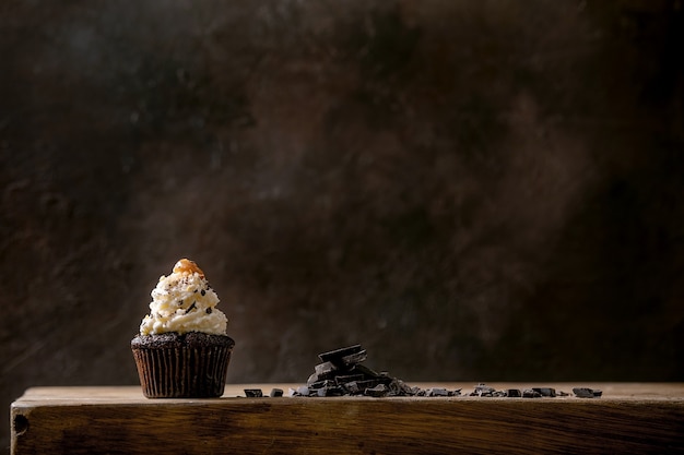 Homemade chocolate cupcakes muffins with white whipped butter cream and salted caramel on ceramic plate, served with chopped dark chocolate on wooden table. Copy space