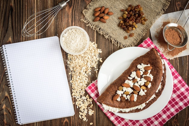 Homemade chocolate oat pancake with cottage cheese on white plate on dark wooden table.