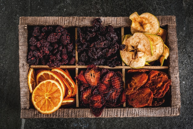 Homemade dried berries and fruits, harvest for the winter: apricots, apples, strawberries, raspberries, cherries, oranges. In old wooden box, on black stone table top view