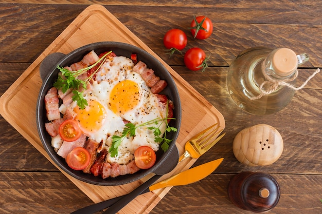 Homemade fried eggs with bacon and tomatoes in a frying pan on wooden background. Classic breakfast. Top view.