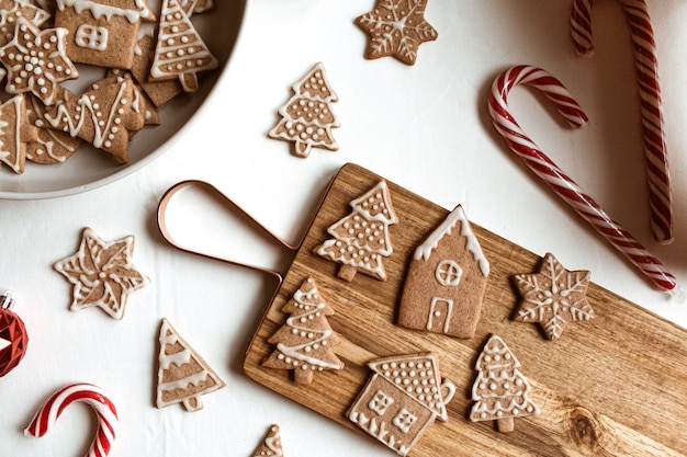 Homemade ginger cookies stars, fir trees, houses on wooden cutting board, stick candies on white. Flat lay, top view Christmas composition.