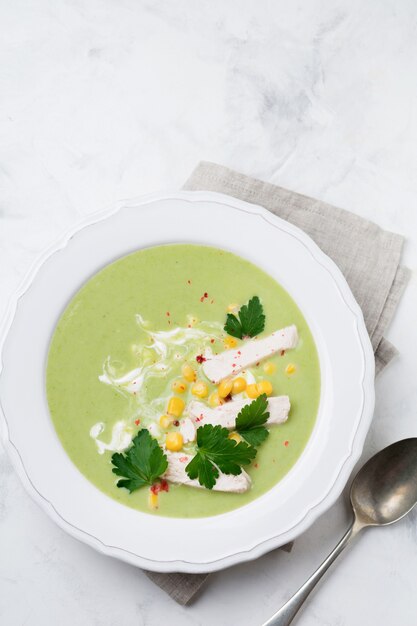 Homemade green soup puree from broccoli, avocado, chicken and corn with cream in rustic ceramic plate on white concrete