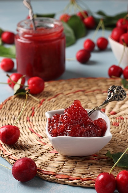 Homemade jam of sweet cherry in a white bowl and jar on a straw rug Closeup Vertical format