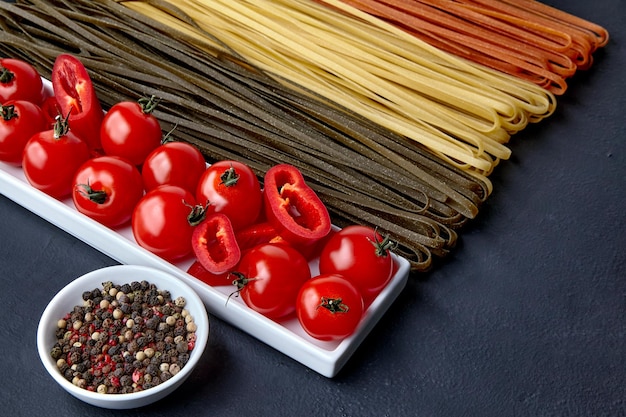 Homemade noodles in different colors from natural herbal ingredients an oblong bowl with tomatoes and red paprika and a bowl with colorful peppers on a black concrete table