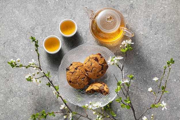 Photo homemade oatmeal cookies on a table with branches of a blossoming tree and green tea in a glass teapot