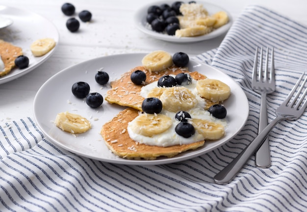 Homemade oatmeal pancakes with yogurt, fresh blueberry and banana at white wooden table.