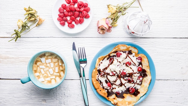 Homemade pancakes or Russian pancakes with chocolate sauce, whipped cream and raspberries on a plate on a white wooden .