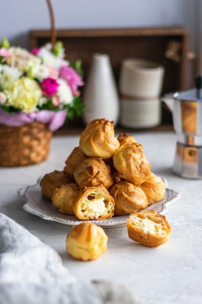 Homemade profiteroles with cream in a gray plate on a white table. Basket with fresh flowers